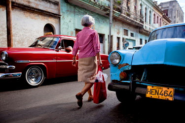 A woman walks past two classic American cars, used as taxis.