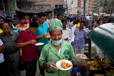 Zafar, surrounded by customers, holds a plate of fried snacks that he cooks and serves from his mobile food cart in the Nari Moitree district.