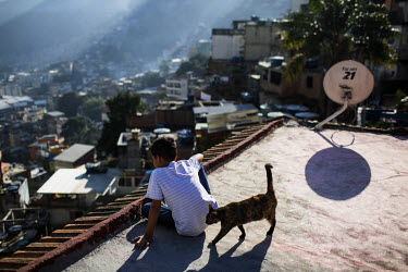 A boy, sitting with a pet cat on a rooftop, looks over Rocinha, the largest single favela in Rio de Janeiro.