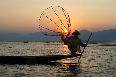 A leg-rowing fisherman with a traditional conical basket net, at sunset on Inle Lake. Most of the lake's fishermen struggle to make a living from fishing and earn more money selling tourists the right...