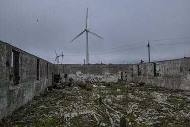 A workshop on Oktyabrsky's sea shore, ruined by coastal erosion. Behind the derelict building rise several sleek wind turbines.   Since the 1970s the sea has been claiming the land, destroying part of...