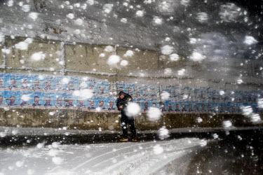 A man walks through a snow storm, past a line of election posters pasted on a wall.