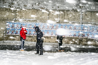 People walking through a snow storm, past a line of election posters pasted on a wall.
