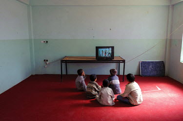 A group of young boys sit on the floor in an orphanage's bare room and watch television.