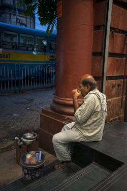 A mobile chai wallah (tea seller) has a rest and drinks a cup of his tea outside a department store.