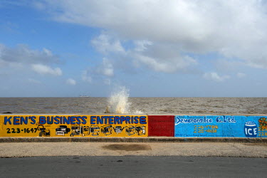 A wave breaks over the sea wall which has hand painted advertisements for local businesses on it.