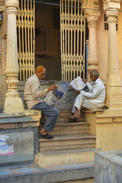 Two elderly men sitting on a stoop reading newspapers.