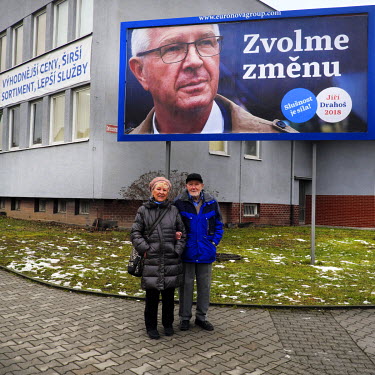An elderly couple, Vera (80, left) and Jara (88) stand beneath a billboard poster advertising their preferred presidential candidate Jirl Drahoo.