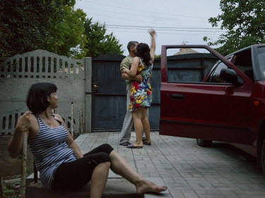 A woman watching her sister dance with her husband outside, beside a car.