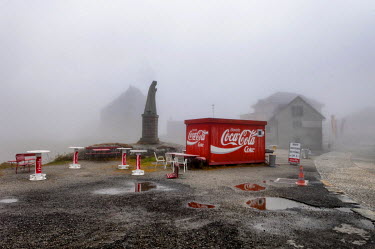 Mist shrouds buildings in a small village where a statue of Sainta Margarita and a stall selling soft drinks stand together at the summit of the St. Gotthard Pass (2,106 m).