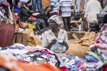 A man sits behind a pile of clothes at an outdoor market selling used textiles.