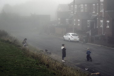 Amanda smokes a cigarette while her children and their friends play in the streets of Easington Colliery on the edge of a green patch of land where the former coal mine stood.