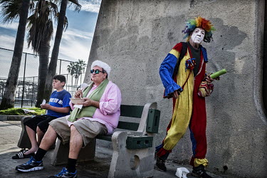 A father and his son eating on a bench in Vencie Beach as a clown street performer passes behind them.