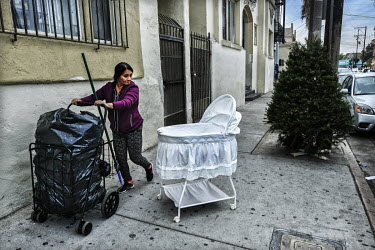 A Latino cleaner passes a baby's cot abandoned on the pavement..