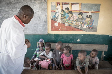A medical worker at a health centre talks with a group of malnourished children who are sitting beneath a picture depicting a group of children eating a meal.