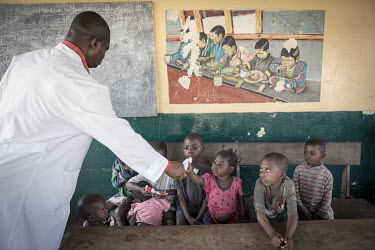 A medical worker at a health centre distributes food to a group of malnourished children who are sitting beneath a picture depicting a group of children eating a meal.