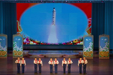 A group of children perform on stage in front of a giant screen showing a ballistic missile blasting off into the air at the Mangyongdae Children's Palace, a centre for extra-curricular learning.