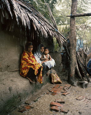 Two women and a child shelter under the roof of a mud hut during a monsoon rain.