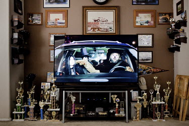 Car racing trophies belonging to Dan Olszewski standing beside a televison playing a commercial. Olszewski is a hero of vintage car racing, who often races at at the Tucson Dragway.