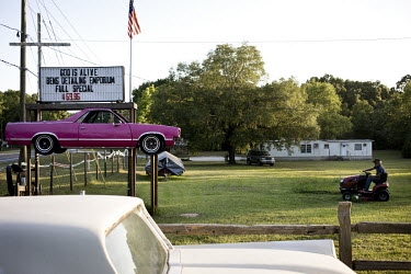 A man mowing a grass lawn at the Bens Detailing Emporium, a car tuning centre advertised by a pink vehicle raised on wooden poles.
