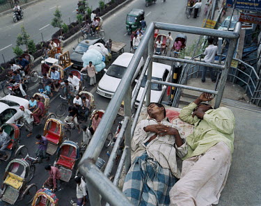 Two men sleep on a pedestrian bridge over looking a busy road crowded with rickshaws and cars.