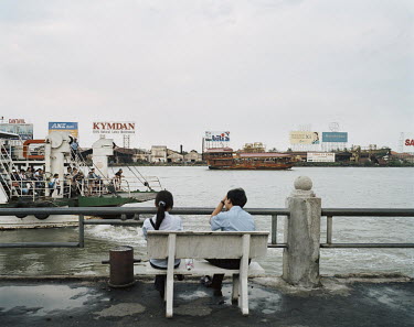 A young couple sit on a bench beside a river as a ferry passes them heading to the dock.