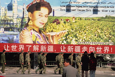 A group of PLA soldiers walking past a Communist Party billboard advertisment featuring a Uyghur woman and promoting the province of Xinjiang, written in Chinese, Arabic, English and Russian.