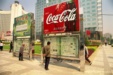 People reading newspapers displayed below a Coca-Cola advertisement in the city centre.