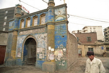 A Uyghur man passes a small mosque surrounded by urban development.