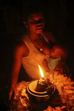 A woman breastfeeds a baby while running her food stall, which is lit by an oil lamp.