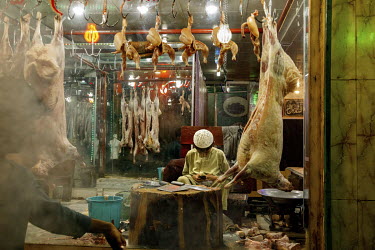 A young boy checks a mobile phone while sitting, surrounded by meat, inside a butcher's shop in Shahr-e Naw.