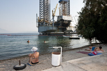 People relax on a beach near a damaged offshore platform moored in the town's the shipyard in the Bay of Kotor.
