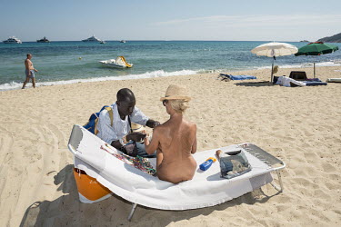 A Senegalese migrant sells a bracelet to a naked woman on a naturist stretch of Pampelonne Beach.