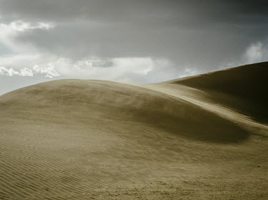 Strong winds blow across a sand dune. A few tufts of grass in the foreground mark the edge of the grassland and where desert begins. Desertification in Northern China is caused by overgrazing, excessi...