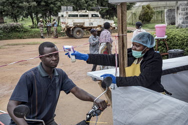 A woman checks the temperature of a man arriving at the local hospital in order to check for people with Ebola infection. In the background a UN peacekeeping force with an armoured vehicle keep watch.