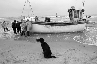 A curious dog watches as fishermen push their boat into the sea, with help of a sea rescue squad in a Zodiak boat pulling it by rope. While on the water, the fishing boats engine got clogged with die...