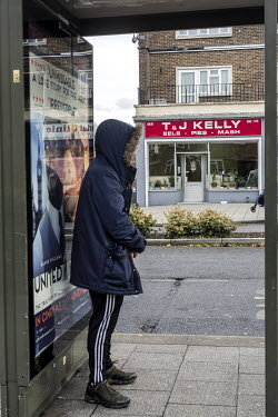 A young man stands in a bus stop opposite T & J Kelly Eel, Pie and Mash shop in Loughton, Essex.