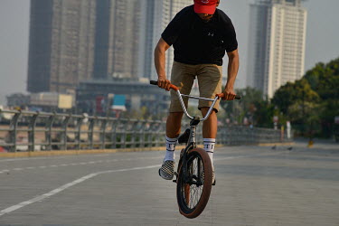 A youth riding a BMX bike practises a move along the Phnom Penh riverfront, with the looming towers of Diamond Island (Koh Pich) in the background.