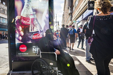 A Muslim woman sits in a wheelchair making a call on a smartphone. Behind her an advertisement on a bus stop promotes Coca-Cola.