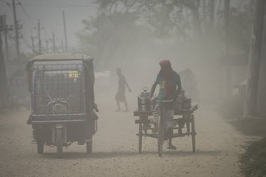 Dust rises from traffic as a cycle rickshaw loaded with metal containers makes its way along an unpaved road.