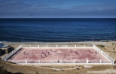 Youths play football on a concrete pitch on the shores of the Mediterranean.