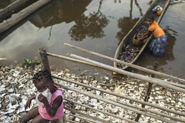 A girl drinks water from a plastci bag used as a container while below a woman hoists a basket full of clams from a boat onto the banks of one of the many water channels that run among the mangrove fo...