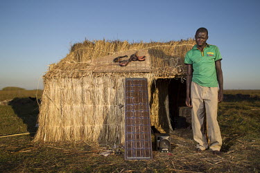 A man stands outside his tempoary grass shelter, beside a solar panel which provides him with electricity and some catfish drying on the roof. During the dry season, people come for a few months to fi...