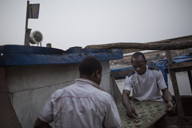 Two men playing 'dam', a popular board game in Africa, on the deck of a disused ship lying in a harbour littered with disused and unfinished vessels. Such wrecks often hamper navigation on the river w...