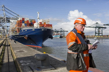 A dock worker looks at a document (the bill of lading) at the port of Antwerp. In the background, the load of a large container ship is visible.