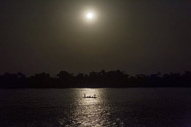 A canoe (pirogue) makes its way along the Congo River, effortlessly by the light of a full moon, flanked by the dark, looming rainforest.