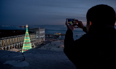 A tourist takes a photo at sunset of the illuninated Christmas tree in the Praca do Comercio with the River Tagus in the background.