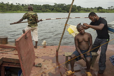 On the deck of a barge sailing up the Congo River a crew member measures the depth of the river with a pole another gets a haircut.