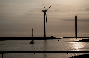 A wind turbine near the harbour basin of the Oosterschelde with a sailing yacht navigating to the open North Sea at sunset.