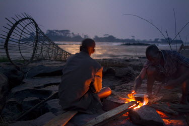 Wagenia fisherman, a bamboo fish trap nearby, cooking on an open fire beside the rapids of Boyoma Falls.